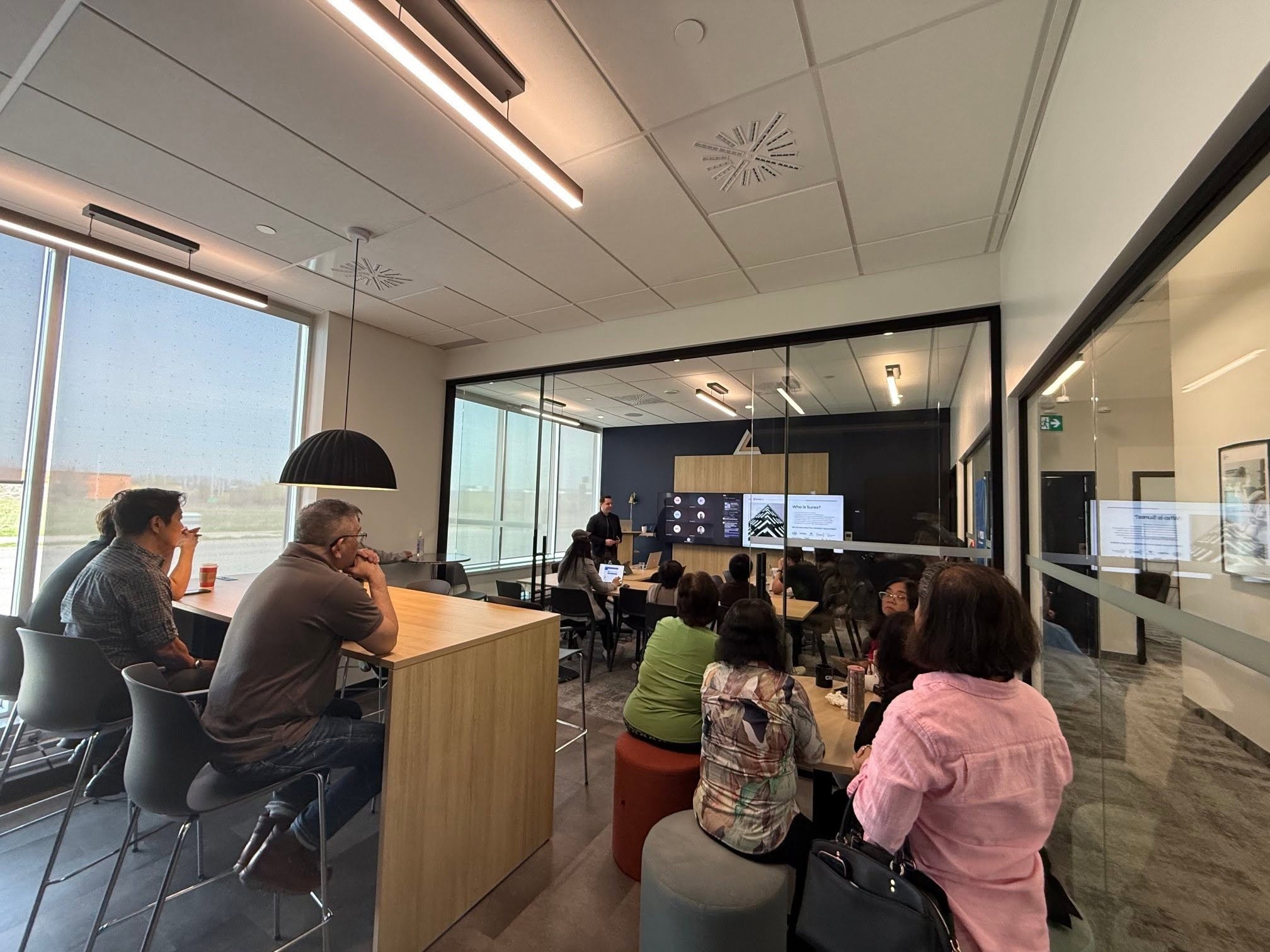 People seated around a conference table watching a presentation in a glass-walled meeting room with ceiling lights and large windows to the left.