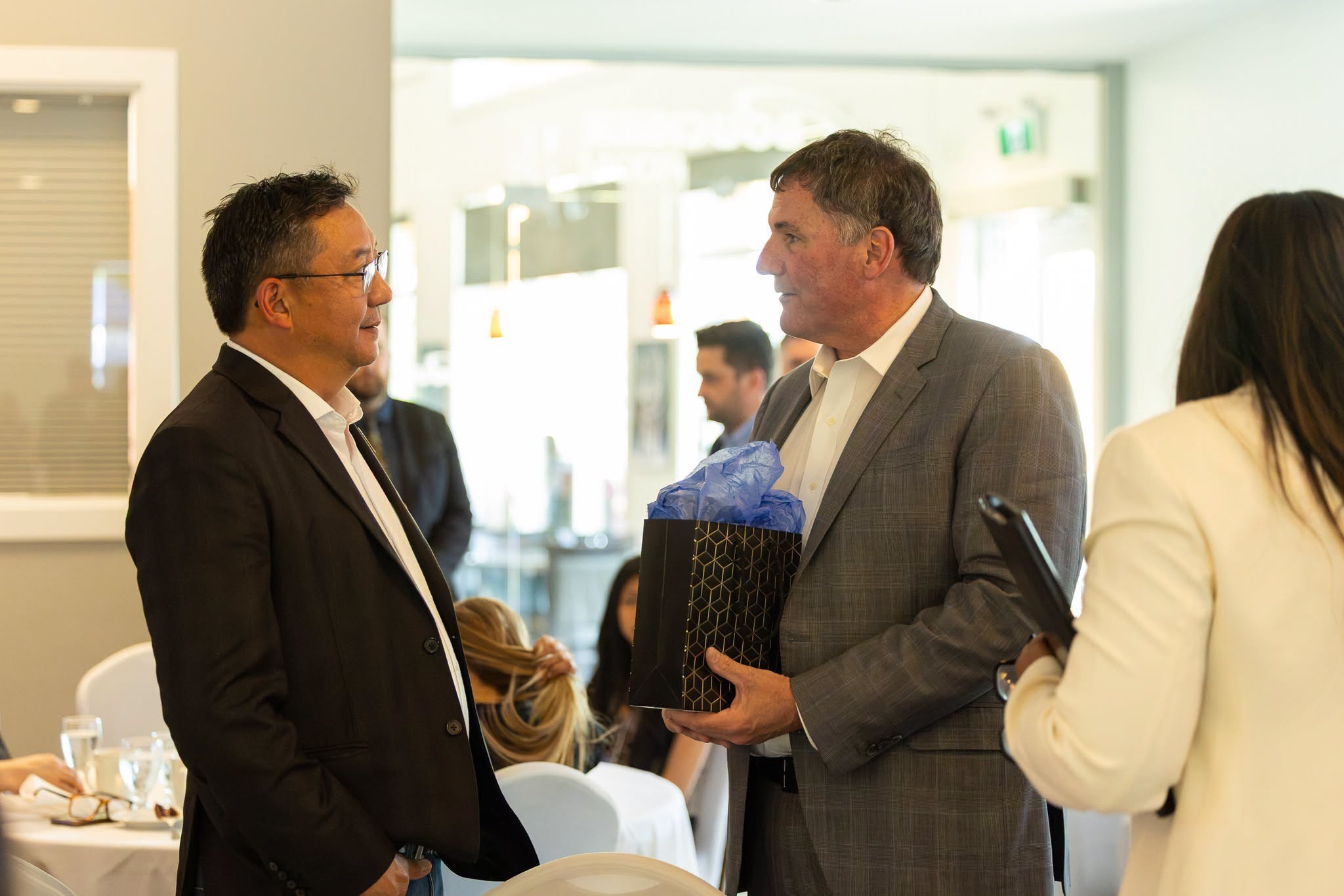 6 - The Canada Times Two men in business attire converse at a reception, one holding a black gift bag with blue tissue. Technicians in background and attendees seated at tables visible behind them.