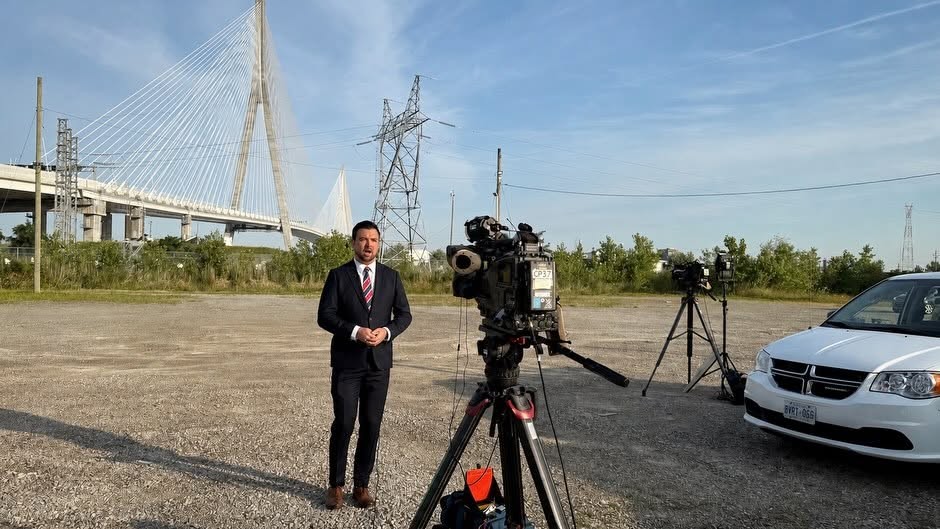 Male reporter in a suit stands beside a camera on a tripod in an open lot, with a bridge and power lines in the background.