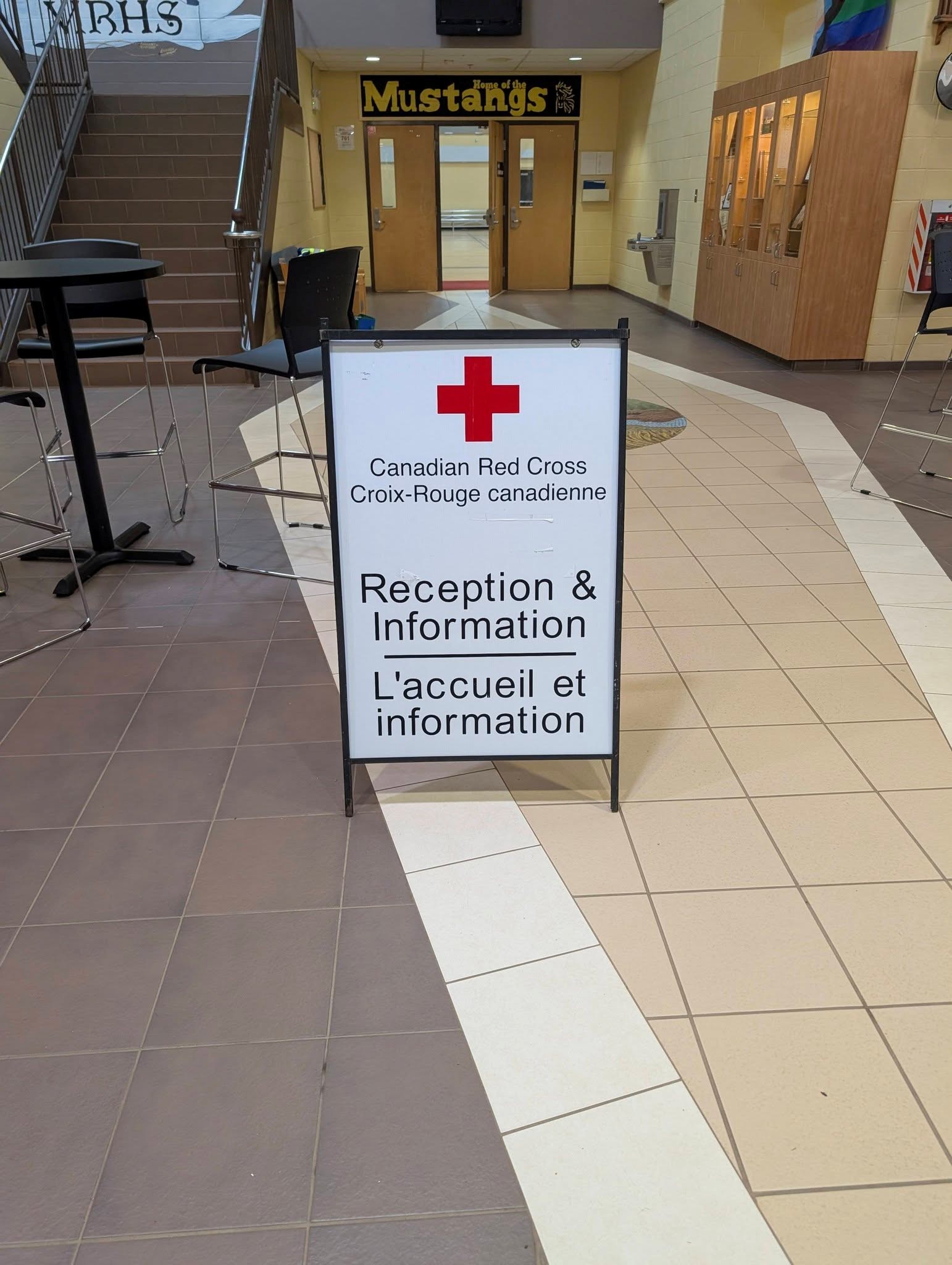 7 - The Canada Times Canadian Red Cross information sign in a school lobby, with double doors labeled 'Mustangs' in the background of the hallway behind.