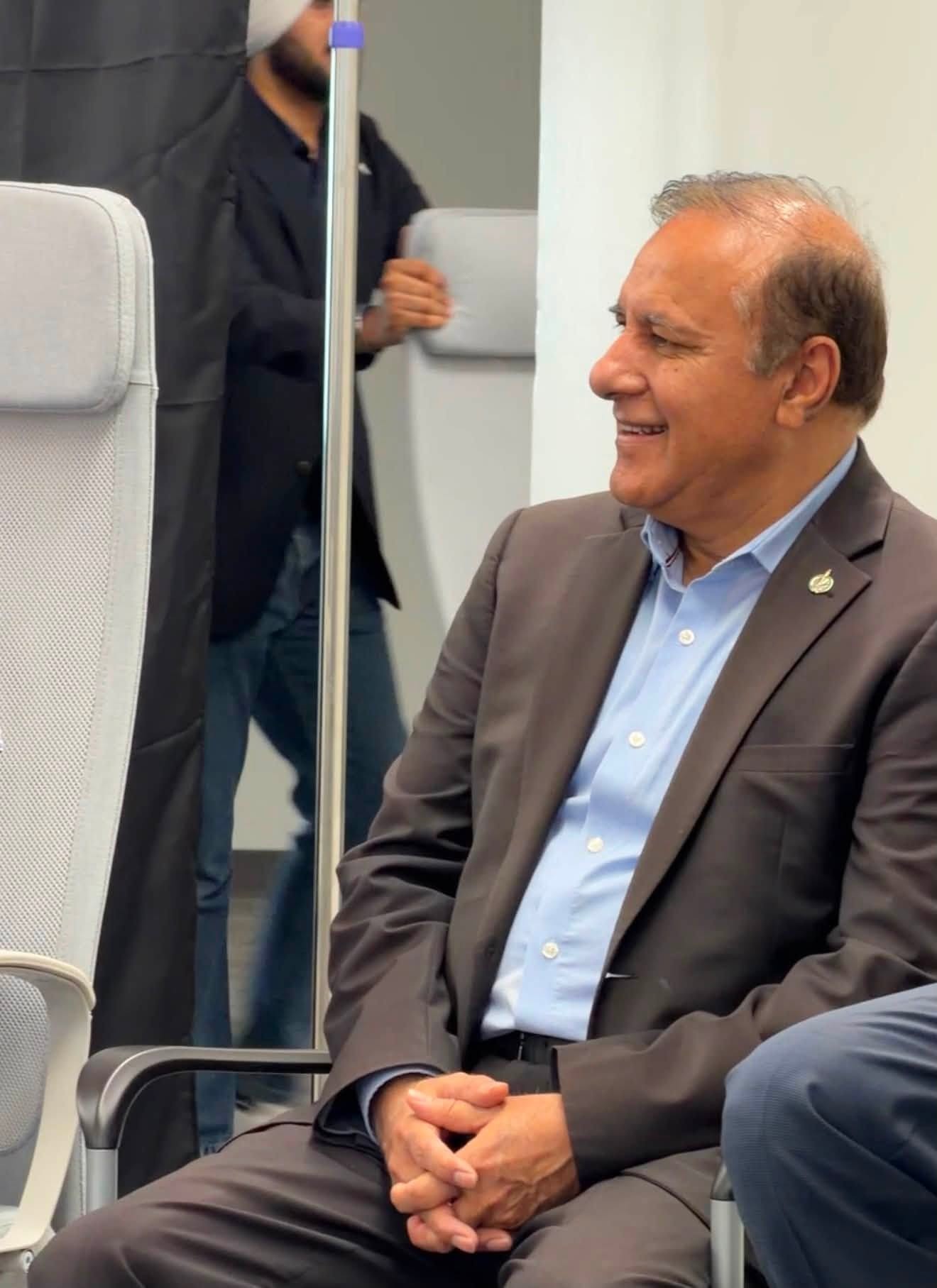 Smiling man in a gray suit and light blue shirt seated on a train, hands folded in his lap.