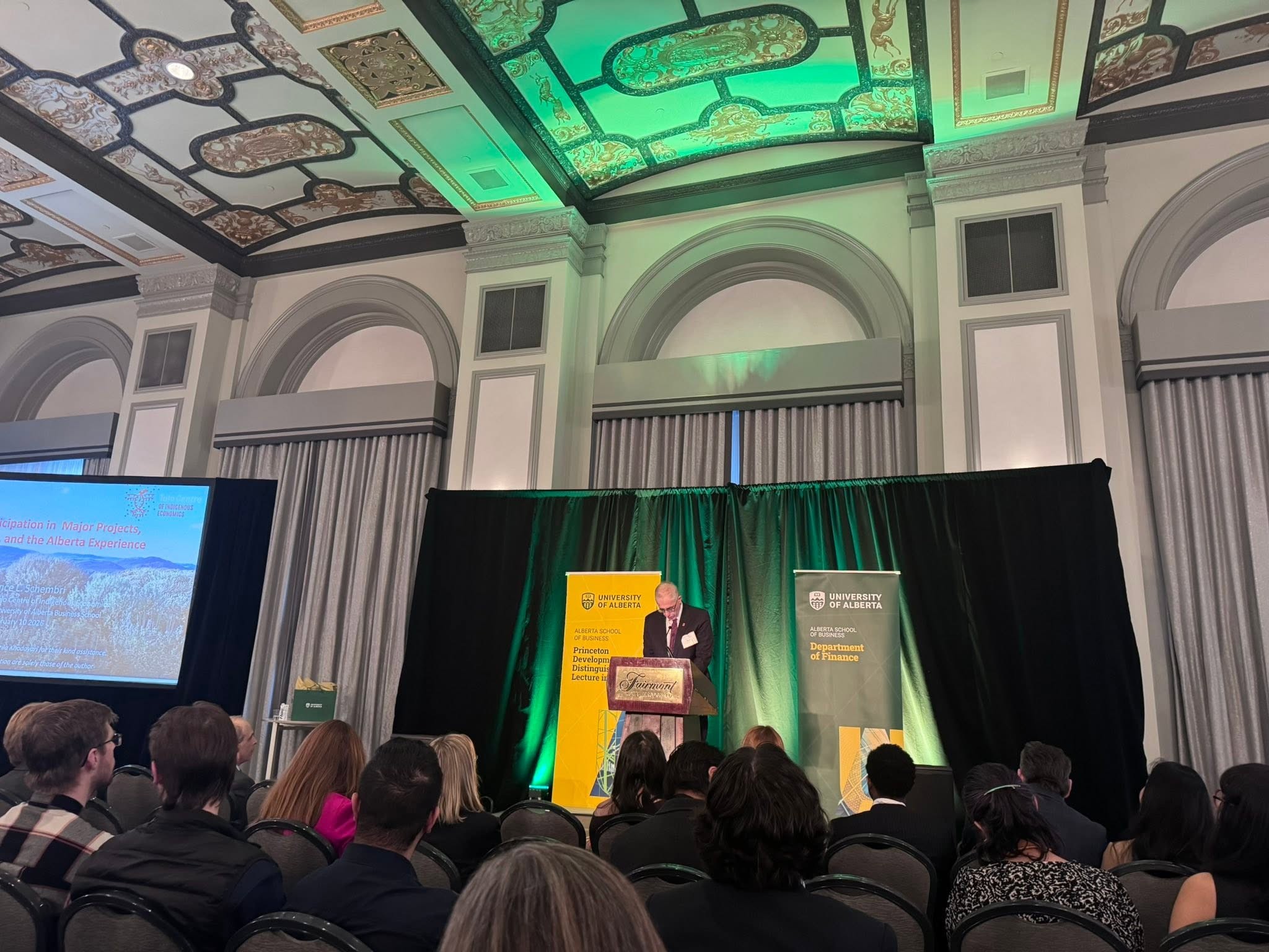 8 - The Canada Times Audience seated in a large conference hall watching a speaker at a podium on a stage with University of Alberta banners and a green-lit backdrop.