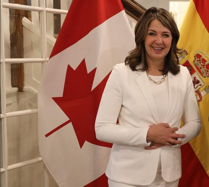 8 - The Canada Times Smiling woman in a white blazer posing with Canadian flag and Spanish flag in the background.