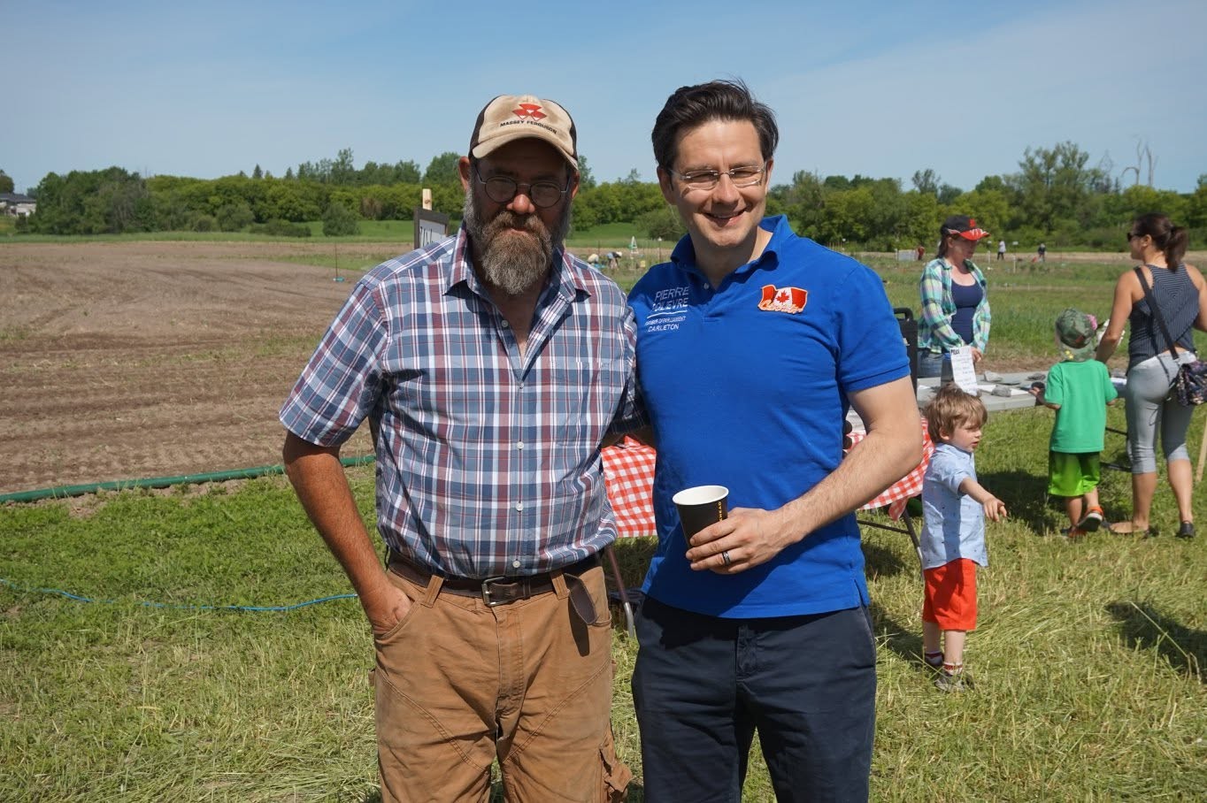 Two men stand together outdoors at a sunny field event, smiling for the camera; one wears a plaid shirt and cap, the other a blue polo and glasses, holds a cup.