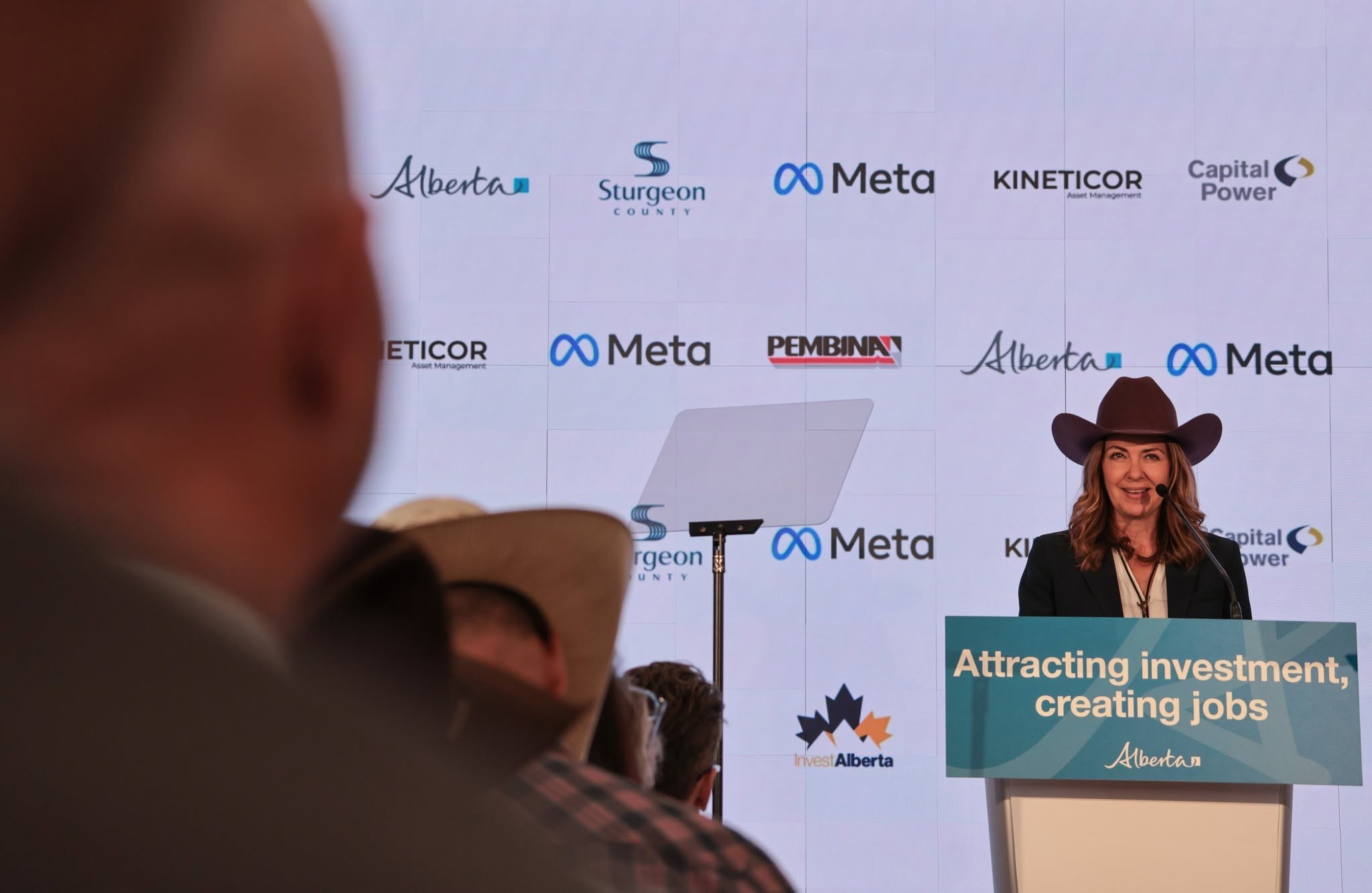 9 - The Canada Times Speaker at a podium delivering a talk, backdrop shows Alberta investment logos and 'Invest Alberta' branding on a large screen behind.