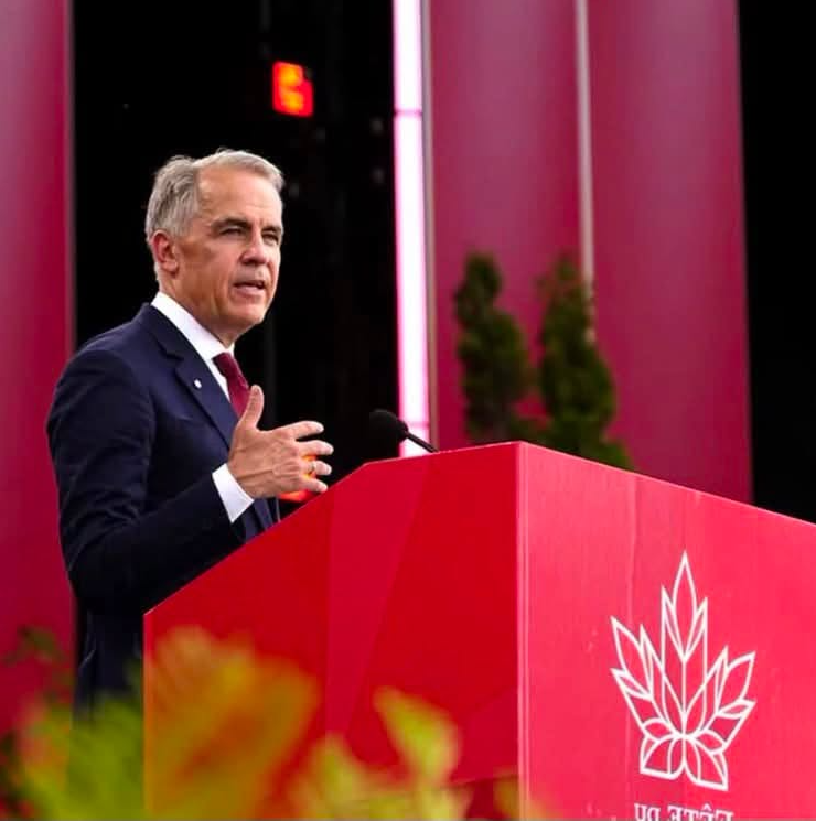 9 - The Canada Times Man in a navy suit delivering a speech at a red podium with a white leaf emblem, in front of pink columns at a formal event